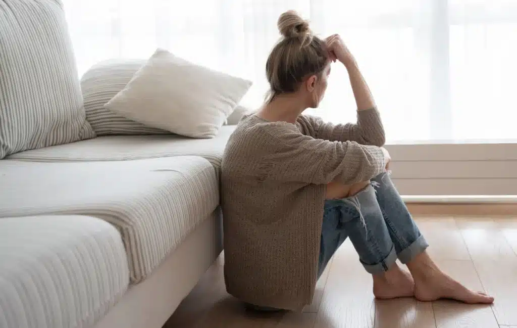 A woman in thought sits on the ground in front of her couch.