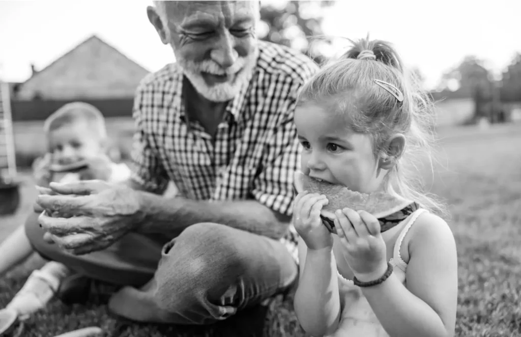 A grandfather sits with his grandkids while the kids eat watermelon.